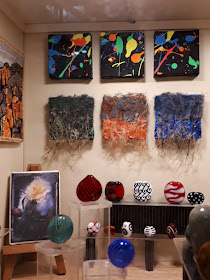 Corner of an art gallery with a variety of colourful tapestry, painted and glass art works on display.