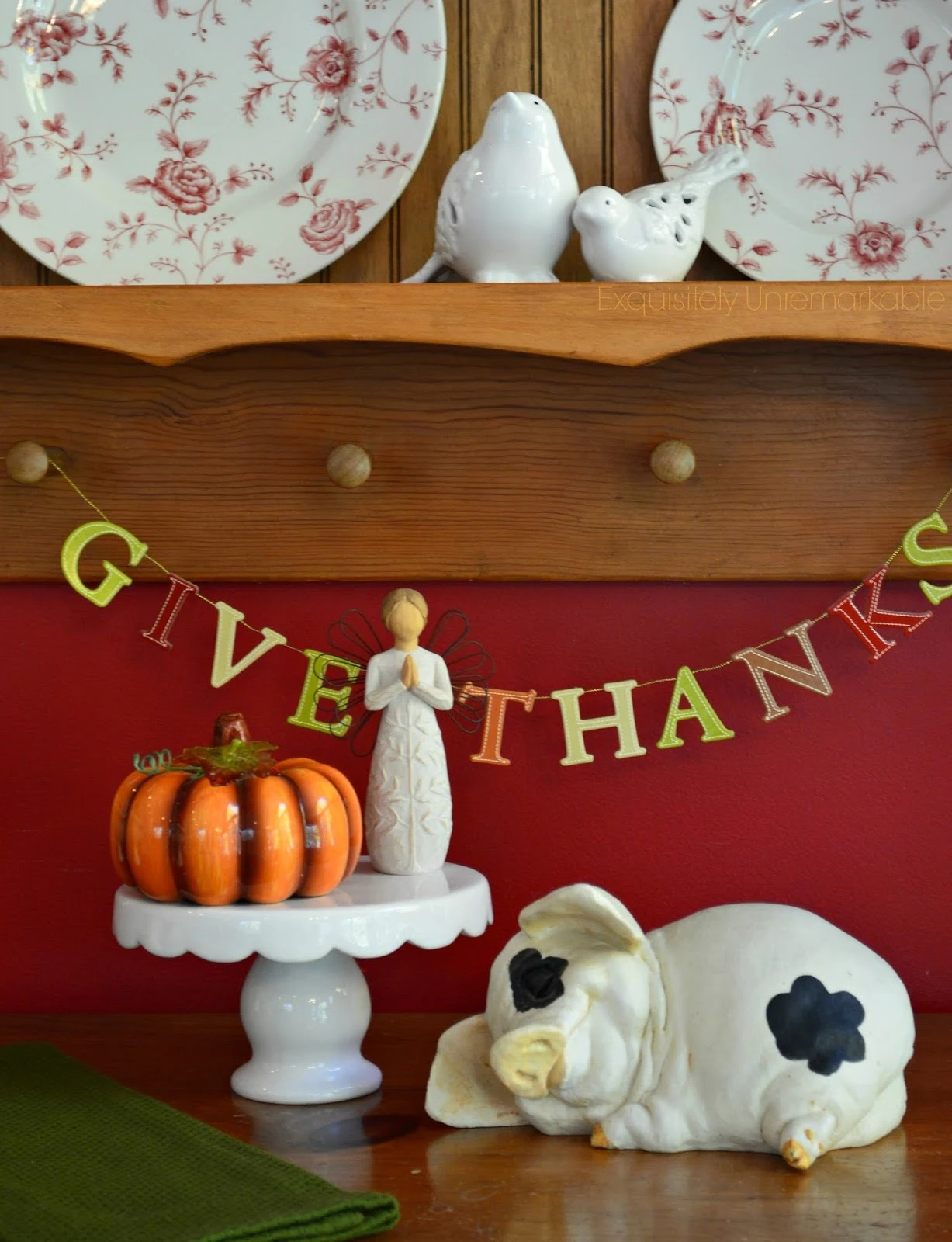 Give Thanks Letter Banner hung on a plate rack next to small statues of angel, pumpkin and sleeping piglet