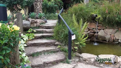 Escada de pedra com pedra moledo tipo chapa com cantos. Escada com pedra com junta de grama com o corrimão de madeira, o lago ornamental com pedra e os pilares de pedra para o lampião no jardim.