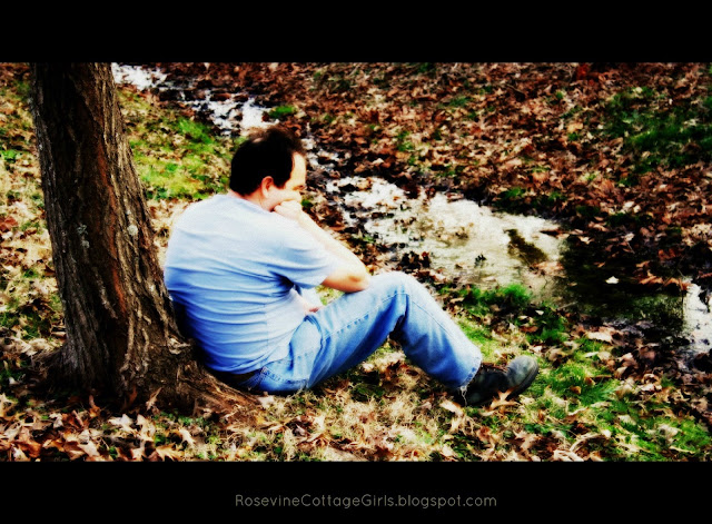 Photo of a man sitting against a tree by a slow moving stream. He has his knees drawn up and is resting his chin on his hand and elbow on his knee. The article is cherishing the tender moments by rosevinecottagegirls.com