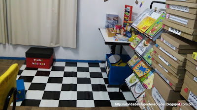 A imagem mostra um tapete de PVC quadriculado, preto e branco. Ao fundo uma caixa vermelha com pinos de encaixe. A direita da imagem uma mesa com vários materiais; uma estante com diversos livros e parte das prateleiras com as caixas de jogos usados no AEE.