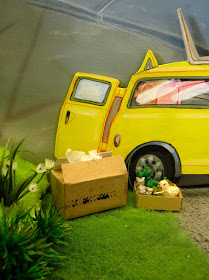One-twelfth scale miniature scene of a yellow Mini van in a garden. It's back doors are open with two cardboard cartons on the ground next to it.