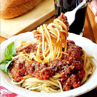 Spaghetti with Slow Cooker Bolognese Sauceon it being twirled on a fork in a white bowl with a red and white checkered napkin on a wooden table with bread and a glass of wine.