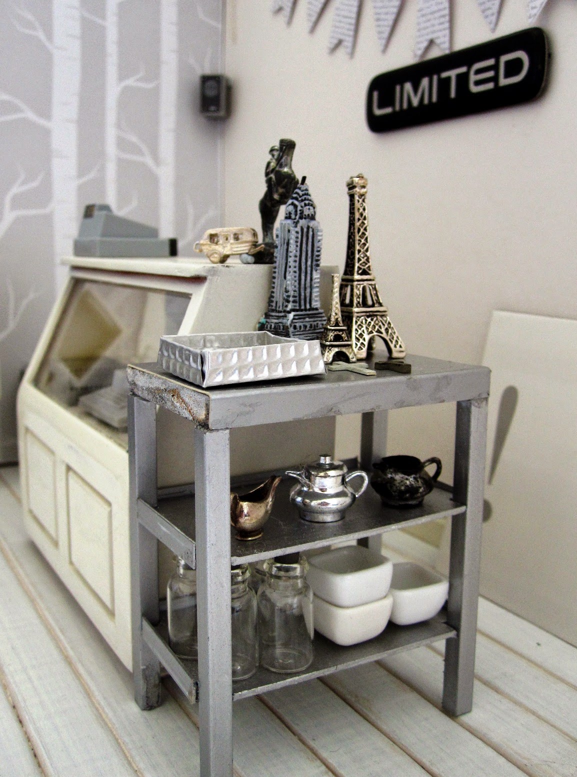 Counter area of a modern dolls' house miniature homeware shop in grey and white. On the counter is a cash register and two silver statues. To the right of the counter is a grey metal shelving unit displaying a selection of model buildings, a range of vintage silver jugs and a number of empty bottles and white bowls.