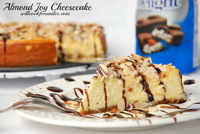 almond joy cheesecake slice on a white plate with chocolate drizzled over it and sliced almonds and the rest of the cheesecake in the background with almond joy creamer to the right 