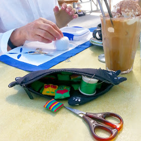 Cafe table with a glass of iced coffee on it. Next to the glass is a cat-shaped pencil case containing miniature cushions, thread and scissors and behind it is a woman working on an appliqué piece.