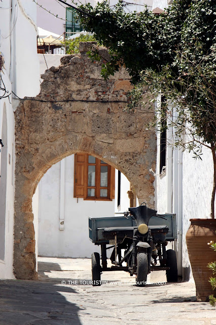 A travel guide to Lindos in Greece. The Touristin. A three-wheeled vehicle on a cobblestoned lane in front of a stone arch.