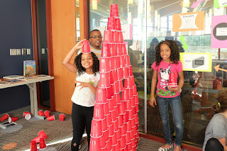 Forbes, Adaora, and Goldie's pyramid structure. Nice hat! A young boy and two girls stand beside a tower made of red plastic cups. The youngest girl wears a cup on her head as a pretend hat.