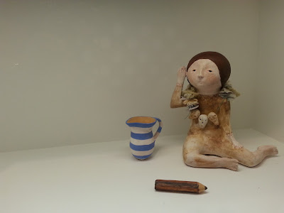 Close-up view of a gallery display of a miniature clay figure, blue and white striped jug and pencil