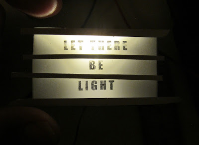 Modern dolls' house miniature light box, lit in the dark with the words 'let there be light' on it.