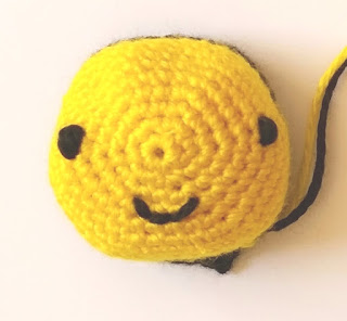 A circular piece of crochet worked in a spiral of double crochet stitches  (USA single crochet) with two black eyes and a smiley mouth embroidered on it. A black thread and a yellow thread can be seen coming from the crocheted piece on the right hand side.