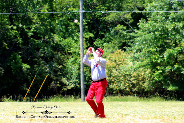 Man playing baseball in a vintage 1860s uniform. The baseball is starting to descend and he is posed to catch the ball double handed. He is wearing red pants, a gray shirt with large "P" on the breast of the shirt with a red baseball cap.  The field is surrounded by large trees. The article is Vintage Baseball by rosevinecottagegirls.com