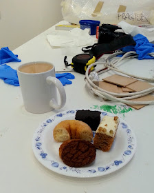 A mug of tea and a plate of cakes and biscuits on a table with tools for a gallery install, including rubber gloves, a tape measure, an extension cord and something wrapped in bubble wrap marked 'fragile'.