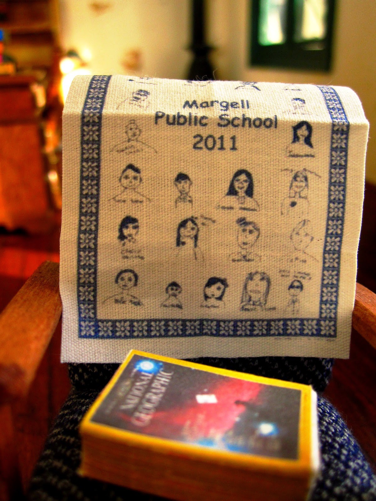 Close up of a 1950s-style miniature chair, displaying a 2011 fund-raising tea towel for Margell Public School and a stack of old National Geographic magazines.