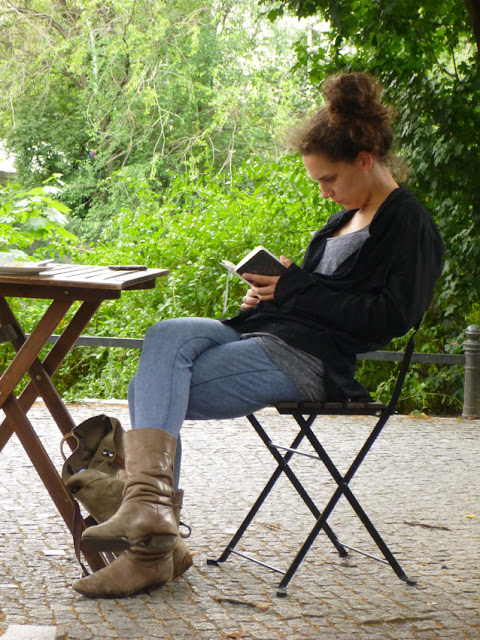 Eine Frau sitzt auf einem Holzklappstuhl an einem Holzklapptisch und liest, hinter ihr ein Weg und viel Parkgrün