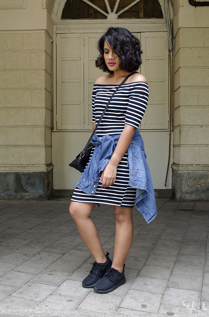 An image of a young woman fashion blogger wearing a striped bodycon river island off shoulder dress, nike rose all black sneakers, denim shirt, accessorize crossbody mini bag and rimmel kate moss pink lipstick with a curly bob hairstyle and pink nails