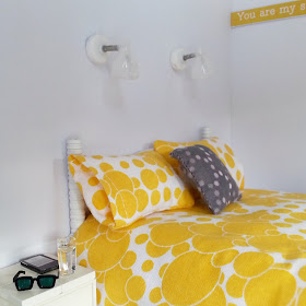 One-twelfth scale modern miniature bed with yellow and white spotty bedding. On the bedside table next to the bed is a Kindle, a glass of water and a pair of sunglasses. Above the bed are two reading lights.