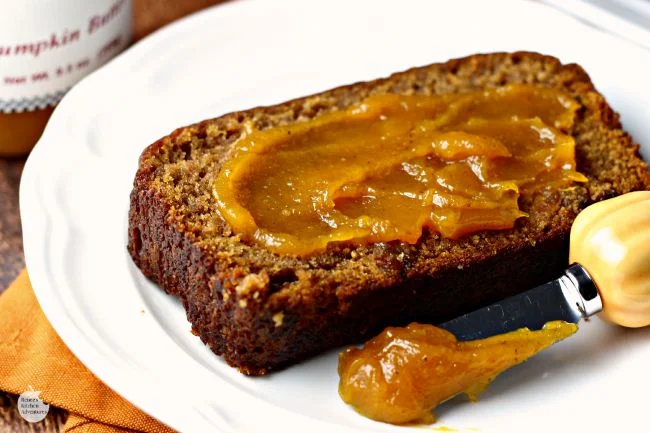 A slice of Spiced Pumpkin Bread by Renee's Kitchen Adventures on a white plate with pumpkin butter spread on it and a small spreader with pumpkin butter on the tip sitting next to the slice