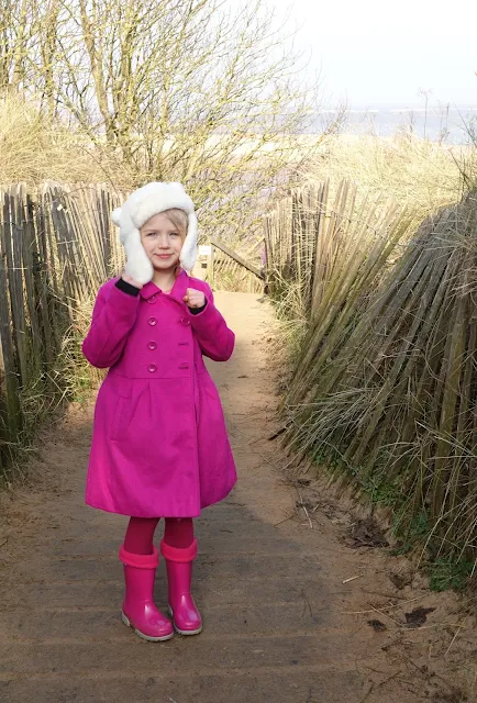A girl in a fluffy white bear hat, pink coat, pink tights and pink wellies on the path leading to the beach at Wells-Next-The-Sea