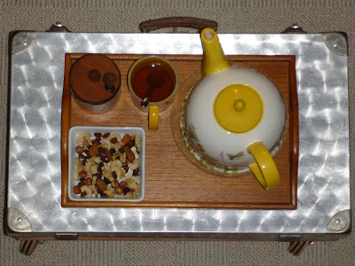 Metallkoffer-"Tisch" mit Holztablett, darauf eine Kaffeekanne, eine Teetasse, Honig und ein Schälchen mit Studentenfutter