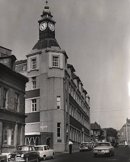 A tall breweries offices building with a clock tower on top. A security guard stands outside the entrance