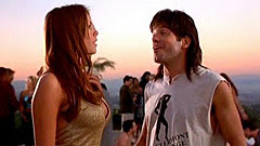 Nikki Cox and David Cross by fire pit Nikki Cox in profile in a tight gold top as the Beer Girl with the sun setting behind her and David Cross as Ronnie in Run Ronnie Run movieloversreviews.filminspector.com