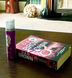 A tall purple and silver bottle with a silver star and Superdrug Extra Firm Hold Hairspray in small black font next to a rectangular book saying dan brown in white font on a brown rectangular table  on a bright background  