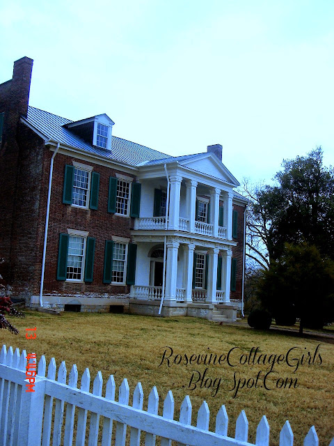 Photo of the Brick front of the Carnton plantation with white pillars on the top and the bottom deck. It has four windows on the top front and four windows on the bottom floor. The bottom center has the front door and the top center has a door to the upper deck. There is a white wooden picket fence around the yard. by Rosevinecottagegirls.com