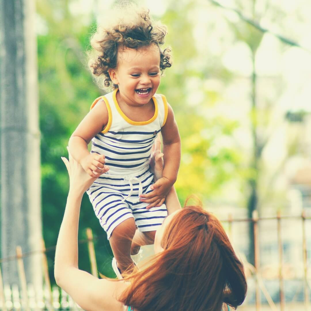 A red headed woman raises her child into the air (her back is to the camera), the child is wearing a blue and white striped t-shirt and shorts combo. He has dark brown curly hair and has a huge smile on his face. There are blurred trees in the background. Image for "Things I've learned about myself since becoming a mother"