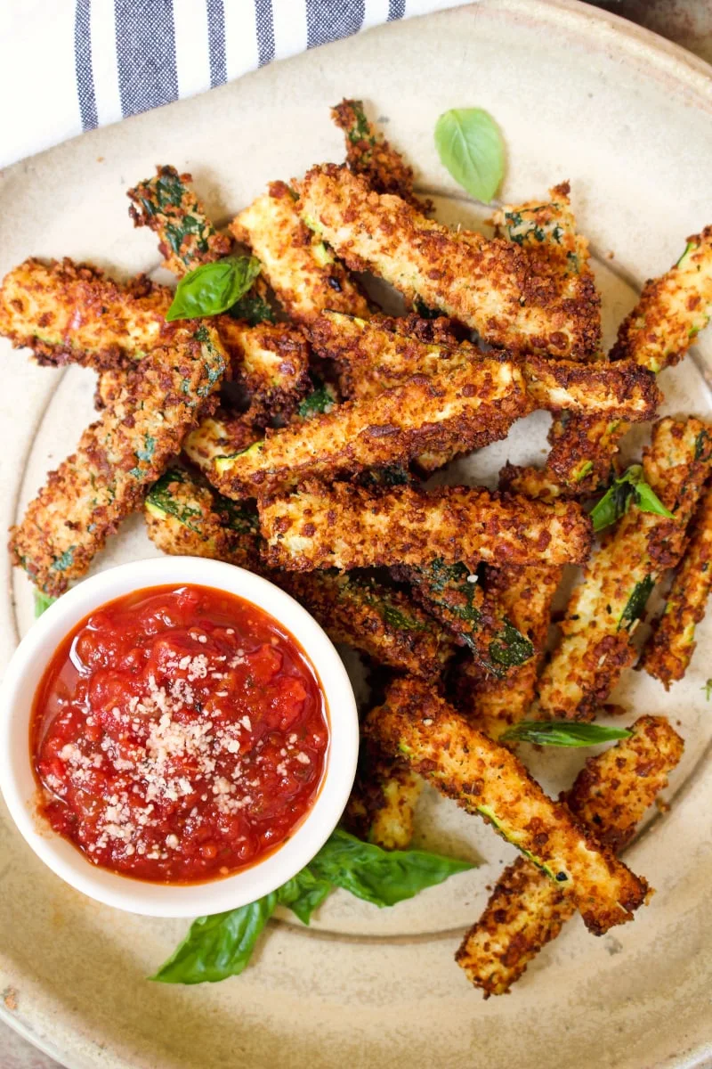 Air Fryer Zucchini Fries coated in a seasoned panko and parmesan cheese coating on a tan plate with a blue and tan striped kitchen towel next to it.