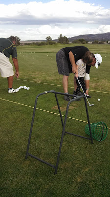 At the golf driving range, Golf coach, Mark helping 5 year old, Jillian, with her foot placement. He is bending over her with his left hand on her left foot.