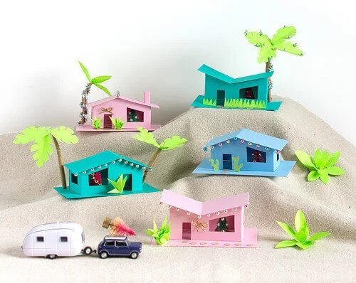Five mid-century ranch houses made of paper with holiday decorations, palm trees, car and trailer