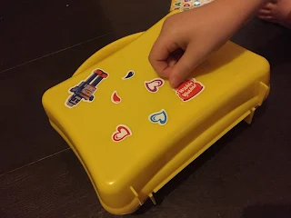 A yellow Hartley's lunchbox being decorated with stickers by a child's hand