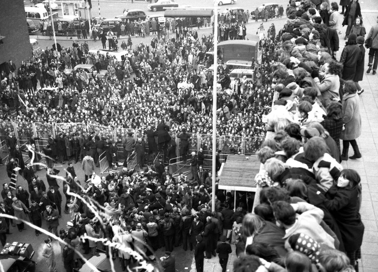 The Beatles (to left of flagpole) are surrounded by newsmen and press photographers and overlooked by hundreds of fans after their arrival at London Airport, England, having flown in from New York on February 22, 1964.