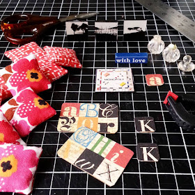 Selection of hand-made one-twelfth scale modern miniatures laid out on a cutting mat with various full-sized tools. Included are cushions, pictures, perfume bottles and place mats.
