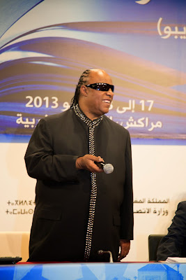 Stevie Wonder standind behind a desk next to a panel of speakers, holding a microphone.