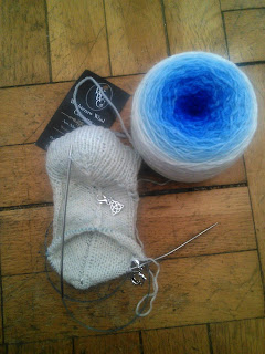 A cuff-down sock on the needles with a gradient cake of yarn next to it.  There are two stitch markers visible on the sock.  The yarn cake is a gradient of white to deep blue. 