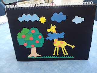 A girafa Gigi está feliz, pertinho da árvore, com a cabeça nas nuvens.