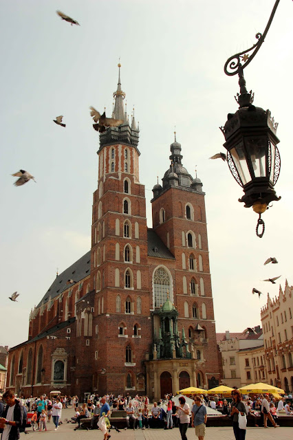 Pigeons flying overhead a crowd of people leisurely strolling in front of a red brick Basilica.