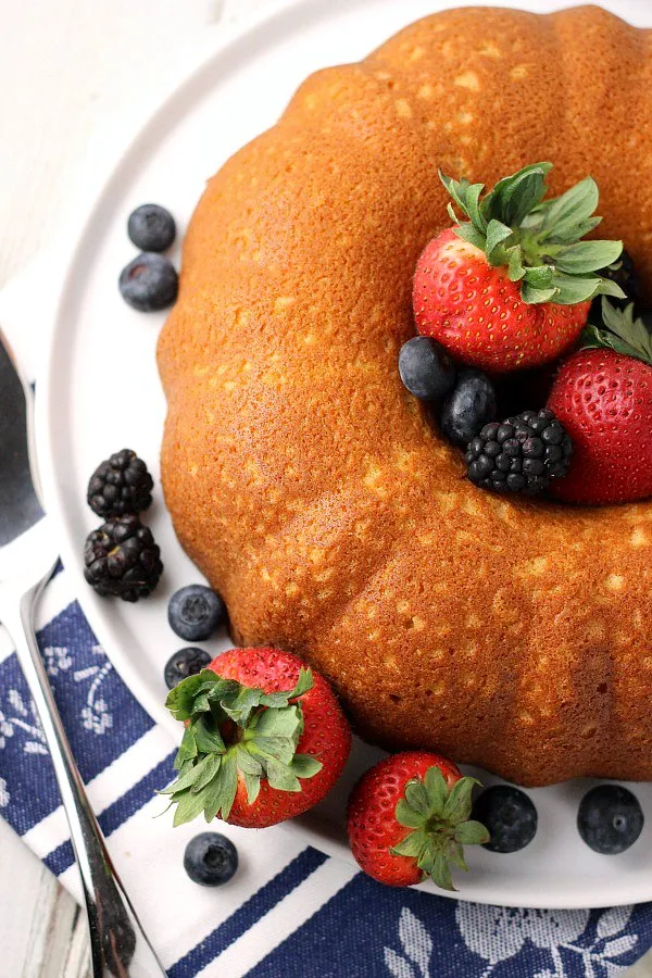 Overhead view of whole Buttermilk Pound Cake on white cake plate, surrounded by blueberries, strawberries, and black berries with server to the side