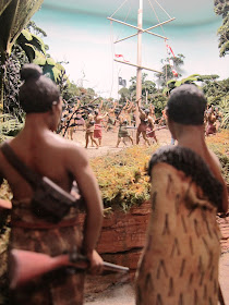 Diorama of 19th-century Maori with muskets on a track in the bush, watching a crowd of people around a flagpole in the distance.