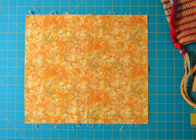 The fabric cut to height and width is laid out flat on the cutting mat. The side edges of the fabric are straight and parallel.  The grid indicates the dimensions of the fabric.  Part of the crochted bag can be seen in the top corner for a sense of scale.