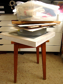 Wooden stool with a pile of wooden and cardboard pieces on top of it, topped with a plastic container full of fabric.