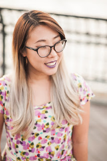 Image of Franny Choi. Image of Franny Choi smiling to the right of the camera. She wears an abstract, floral print dress in shades of pink, yellow, turquoise and black. She wears a lilac shade of lipstick and softly cat-eyed framed glasses. She has long hair that is dark gold hear her scalp and a very light blonde at the ends.