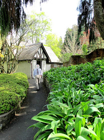 A man walking up a path in an english-style garden to the entry of an arts-and-crafts-style cottage.
