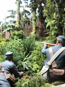 Diorama of 19th-century soldiers with muskets in the bush, aiming at a pa fence.