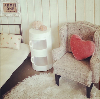 Modern dolls house miniature bedsit scene with a day bed, wing chair, circular fluffy rug and side cupboard in soft shades of white, grey and rose.
