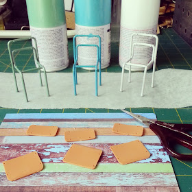 Three one-twelfth scale school chair kits, each painted a different shade of soft blue, green and white, on a cutting mat along with the cans of spray paint and a piece of scrapbooking paper