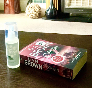 A clear cylindrical tube that says destress mist in black small font with a clear cylindrical lid next to a rectangular book saying dan brown in white font on a brown rectangular table  on a bright background 