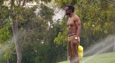 Chidi stands shirtless in the middle of a bunch of sprinklers in a park